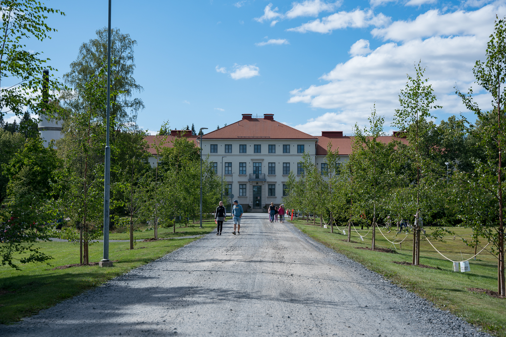 Ihmiset kävelevät hiekkatietä pitkin kohti suurta vaaleaa TB-rakennusta punaisella katolla, ympärillä nuoria puita ja nurmikkoa.